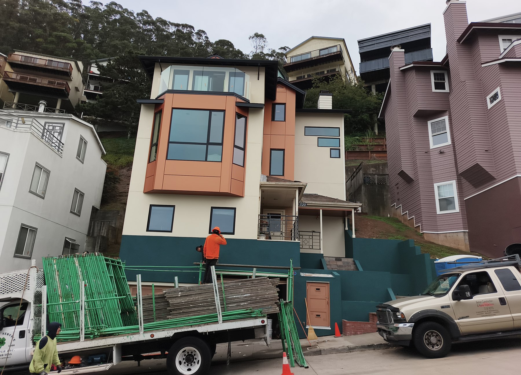Modern hillside home in San Francisco mid-construction with Behan Builders truck on-site, workers on scaffolding, and teal concrete retaining walls in place